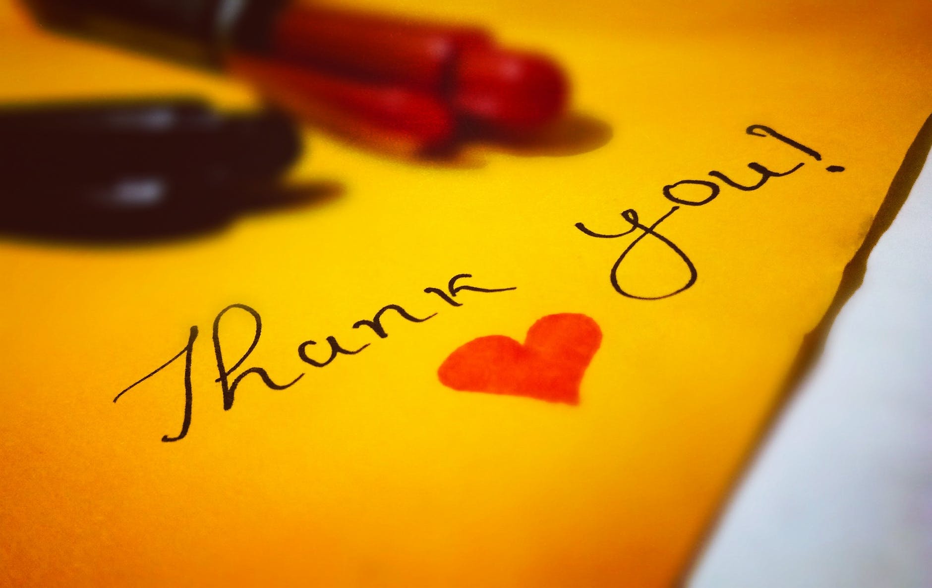 The words Thank You written in cursive on yellow paper with a black pen. There is a red heart drawn below the script, and the pen is resting on the upper part of the page.