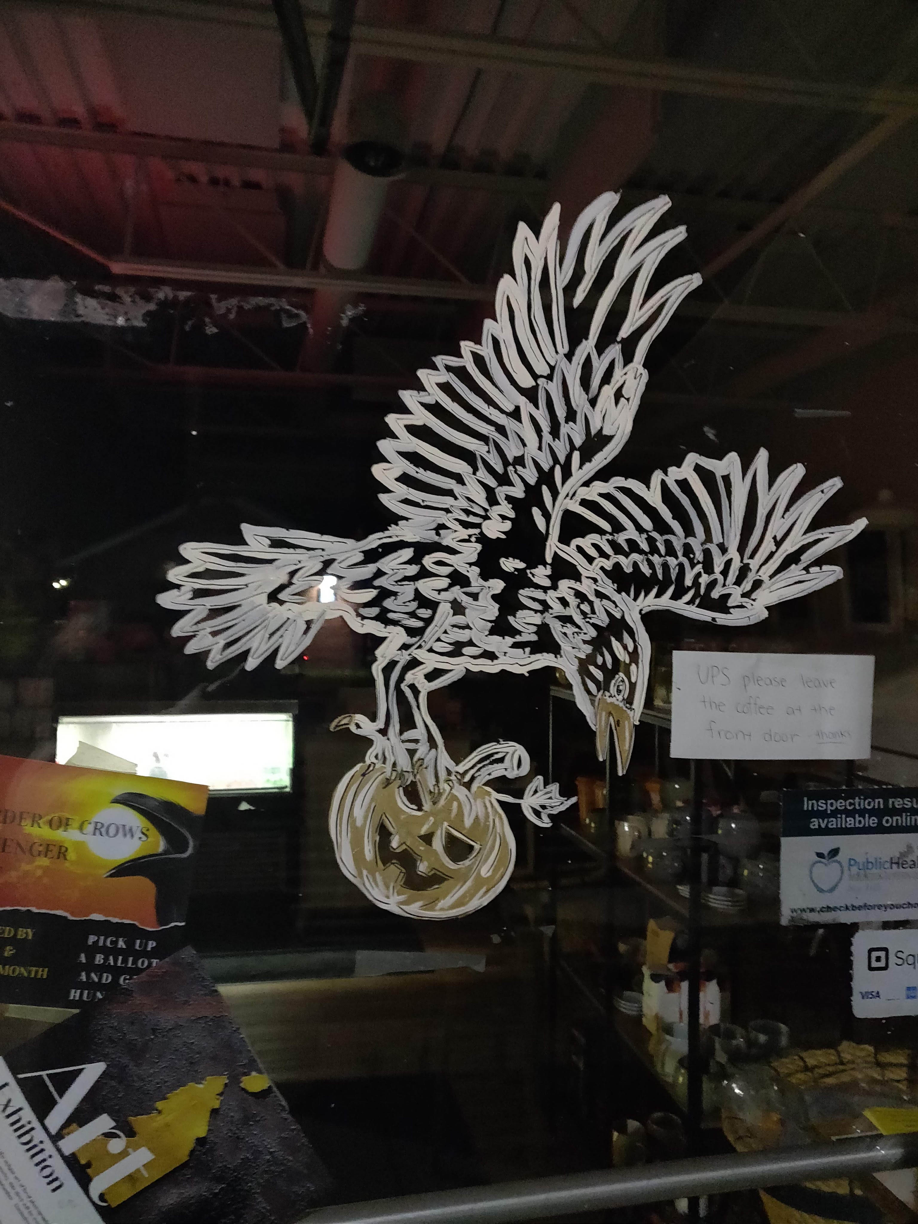 A crow in flight painted on the window of a grocery store, holding a jack-o-lantern in it's talons. 